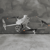 A light gray quadcopter drone with black and orange-tipped propellers, hovering in front of a distressed camouflage metal wall and concrete floor.