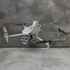 A light gray quadcopter drone with black and orange-tipped propellers, hovering in front of a distressed camouflage metal wall and concrete floor.